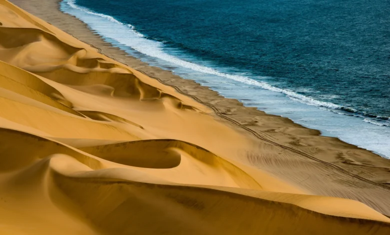 A breathtaking coastal desert landscape showcasing golden sand dunes meeting a deep blue ocean, perfect for Middle East road trips 2024. The image highlights the stark beauty of where desert meets sea, offering a glimpse of the diverse terrains explorers might encounter.