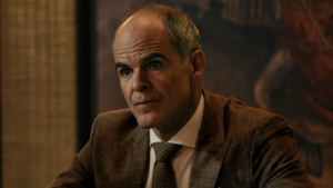 Michael Kelly plays Johnny Vitti, a stern-faced man in a brown suit, radiating intensity.